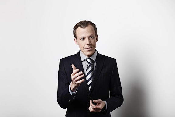 Man wearing a suit and tie, gesturing with one hand as if explaining something