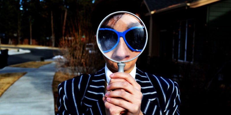 Young man wearing striped suite and sunglasses, whose nose, glasses and forehead are enlarged by magnifying glass he's holding up to his face.